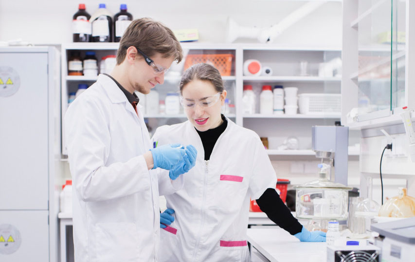 Kaksi tutkijaa laboratoriossa, pukeutuneina valkoisiin takkeihin ja suojalaseihin, tarkastelee näytettä yhdessä työpisteen äärellä.