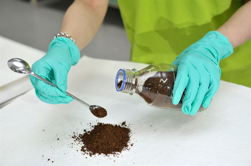 Suojakäsineisiin puetut kädet käsittelevät ruskeaa ligniinijauhetta laboratoriopöydällä