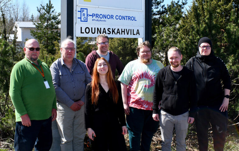 The Pronor Control team standing together outdoors in front of a sign with the company logo on a sunny day.