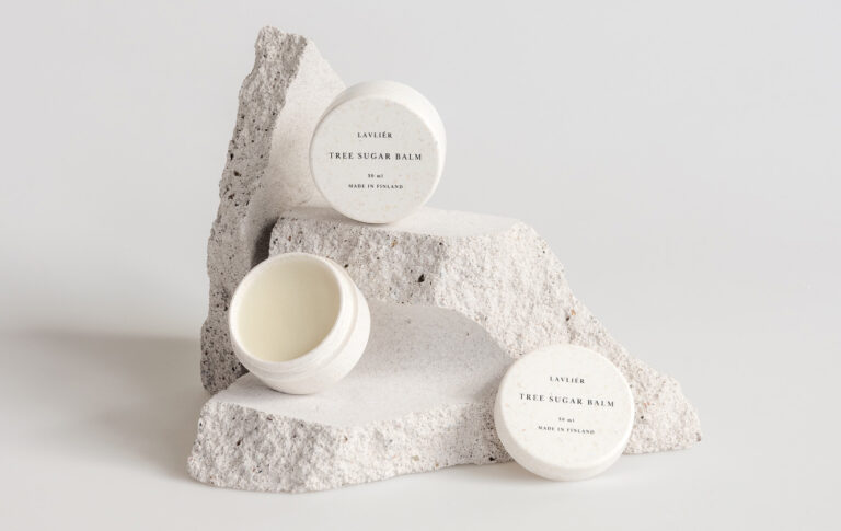 Kolme Lavliér Tree Sugar Balm -voidepurkkia aseteltuna vaaleiden, rosoisten luonnonkivien päälle valkoisella taustalla.