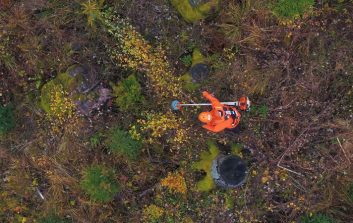 Ilmakuva henkilöstä oransseissa suojavaatteissa tekemässä mittauksia kivikkoisessa metsämaastossa.