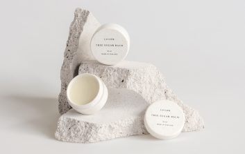 Kolme Lavliér Tree Sugar Balm -voidepurkkia aseteltuna vaaleiden, rosoisten luonnonkivien päälle valkoisella taustalla.