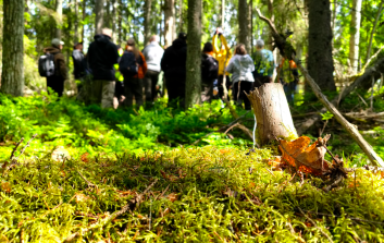 Ryhmä ihmisiä opastetulla luontoretkellä vehreässä suomalaisessa havumetsässä sammaleisen maaston äärellä.