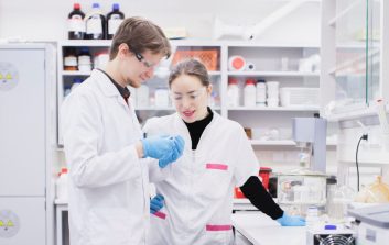 Kaksi tutkijaa laboratoriossa, pukeutuneina valkoisiin takkeihin ja suojalaseihin, tarkastelee näytettä yhdessä työpisteen äärellä.